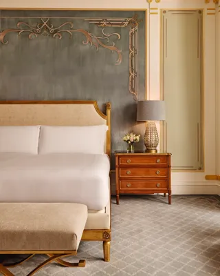 Refined bedroom with a king bed featuring a gold-trimmed headboard, a wooden bedside table with a textured lamp and flowers, and an ornate wall with soft blue and gold detailing.