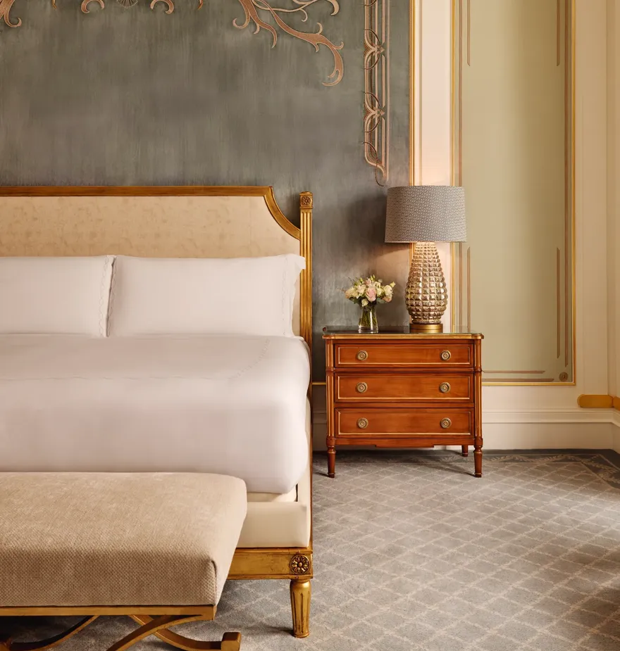 Refined bedroom with a king bed featuring a gold-trimmed headboard, a wooden bedside table with a textured lamp and flowers, and an ornate wall with soft blue and gold detailing.