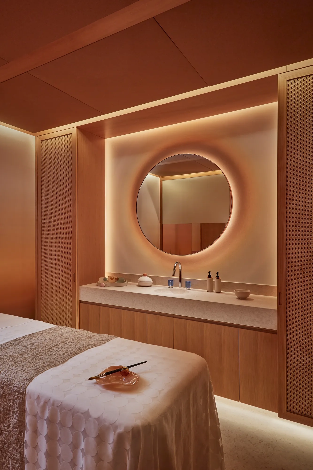 Warmly lit spa treatment room with massage table, round mirror, and natural wood cabinetry for a relaxing experience.