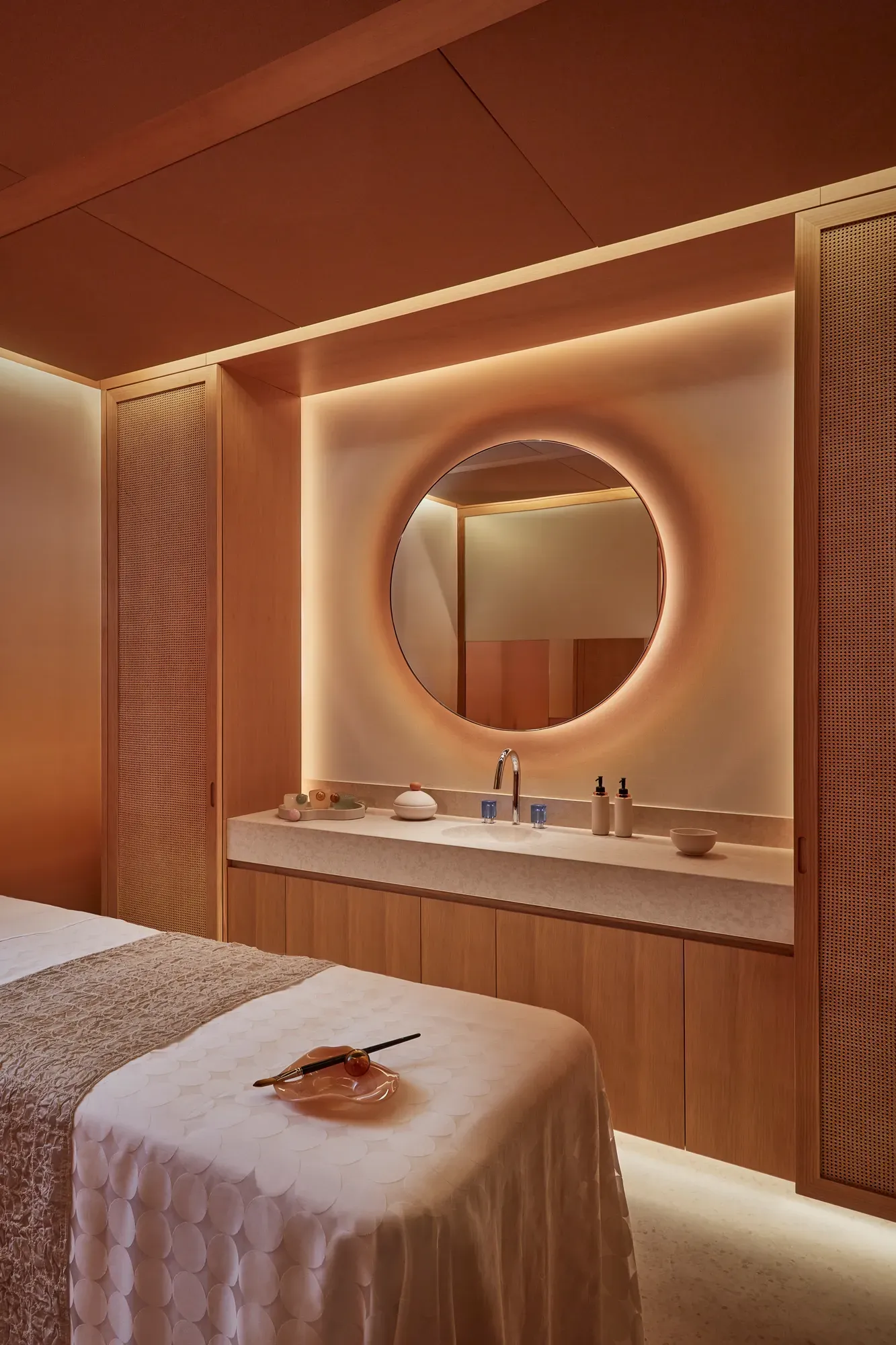 Warmly lit spa treatment room with massage table, round mirror, and natural wood cabinetry for a relaxing experience.