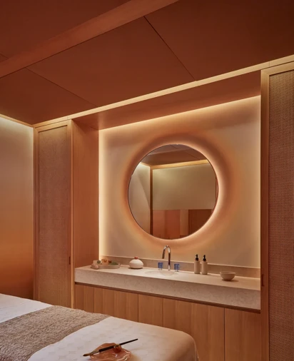 Warmly lit spa treatment room with massage table, round mirror, and natural wood cabinetry for a relaxing experience.