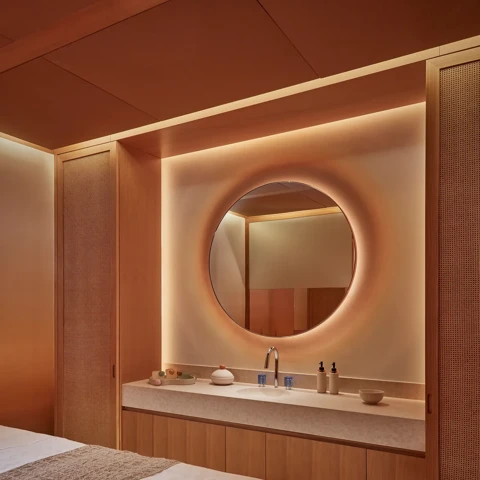 Warmly lit spa treatment room with massage table, round mirror, and natural wood cabinetry for a relaxing experience.
