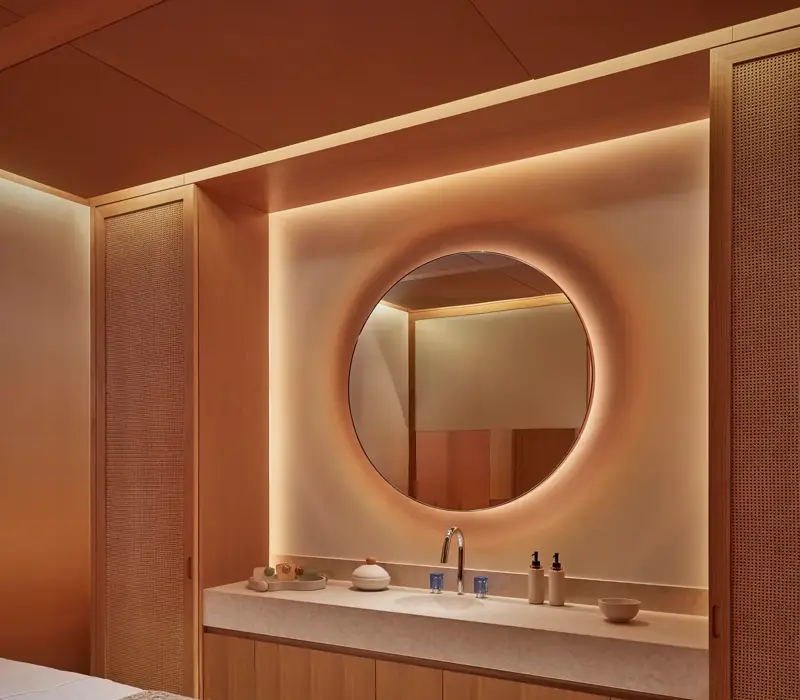 Warmly lit spa treatment room with massage table, round mirror, and natural wood cabinetry for a relaxing experience.