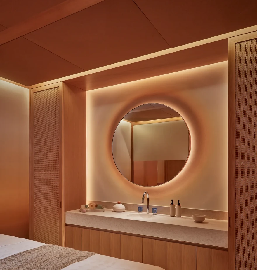 Warmly lit spa treatment room with massage table, round mirror, and natural wood cabinetry for a relaxing experience.