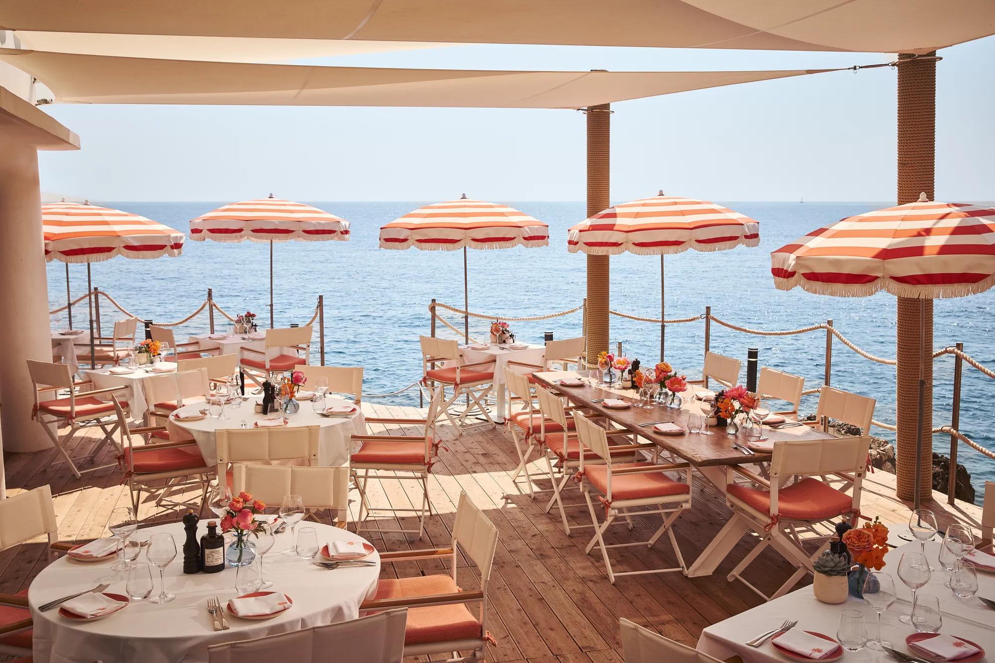 Terrasse de restaurant en bord de mer avec tables rondes et rectangulaires dressées, décorées de fleurs et ombragées par des parasols rayés rouges et blancs, donnant sur la mer bleue calme.