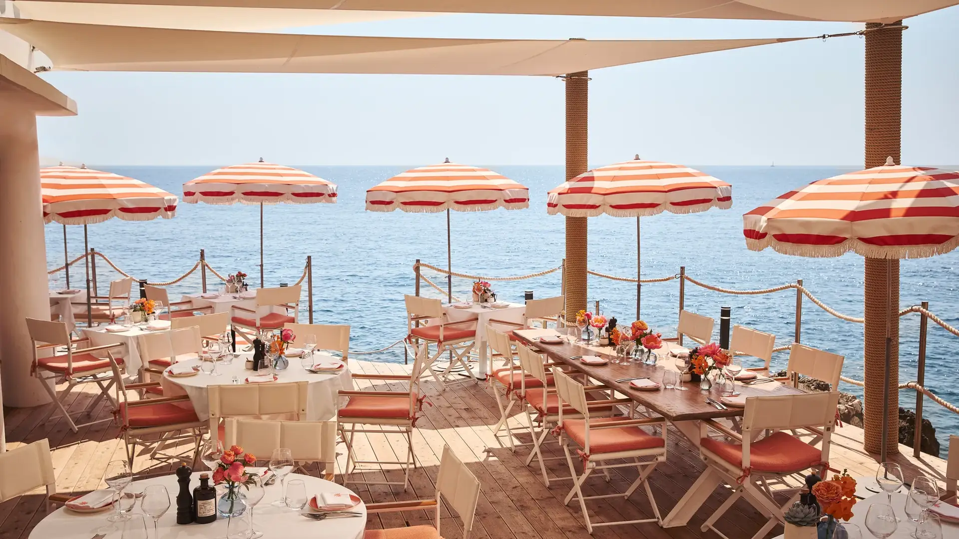 Terrasse de restaurant en bord de mer avec tables rondes et rectangulaires dressées, décorées de fleurs et ombragées par des parasols rayés rouges et blancs, donnant sur la mer bleue calme.