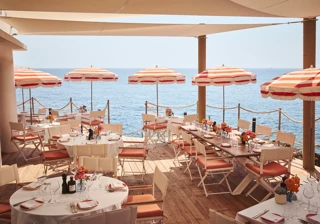 Seaside terrace restaurant with round and rectangular tables set for dining, decorated with flowers and shaded by red-and-white striped umbrellas, overlooking the calm blue sea.