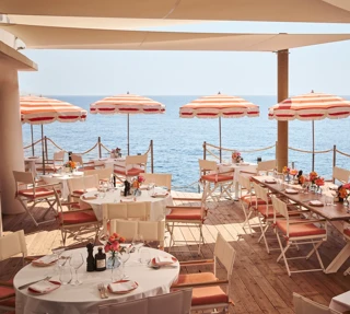 Seaside terrace restaurant with round and rectangular tables set for dining, decorated with flowers and shaded by red-and-white striped umbrellas, overlooking the calm blue sea.