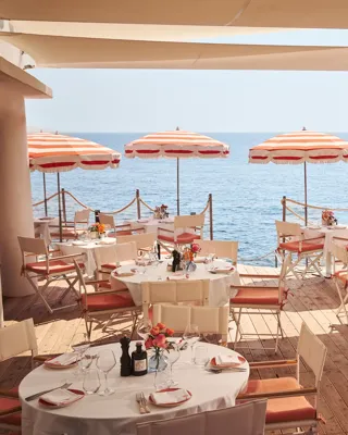 Seaside terrace restaurant with round and rectangular tables set for dining, decorated with flowers and shaded by red-and-white striped umbrellas, overlooking the calm blue sea.