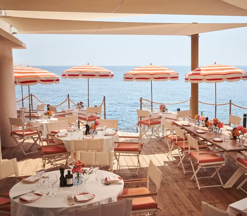 Seaside terrace restaurant with round and rectangular tables set for dining, decorated with flowers and shaded by red-and-white striped umbrellas, overlooking the calm blue sea.