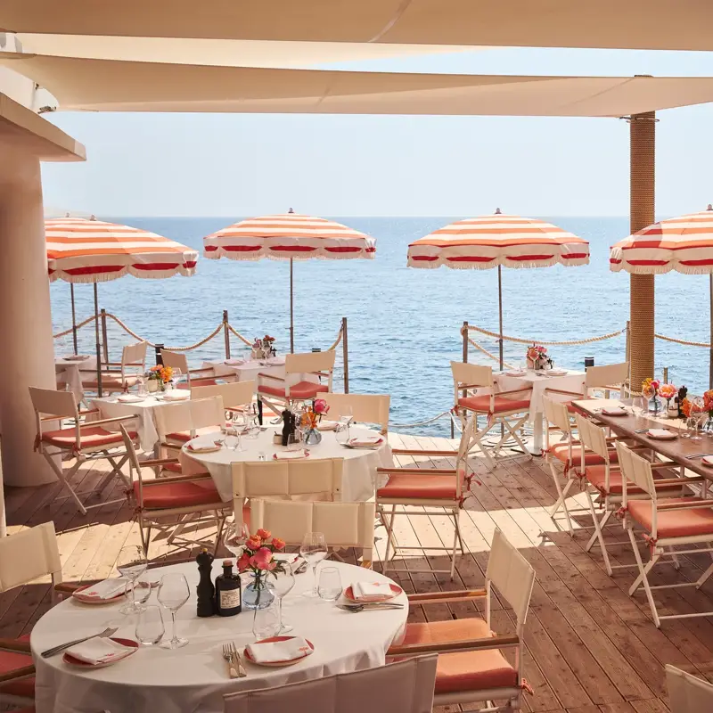 Seaside terrace restaurant with round and rectangular tables set for dining, decorated with flowers and shaded by red-and-white striped umbrellas, overlooking the calm blue sea.