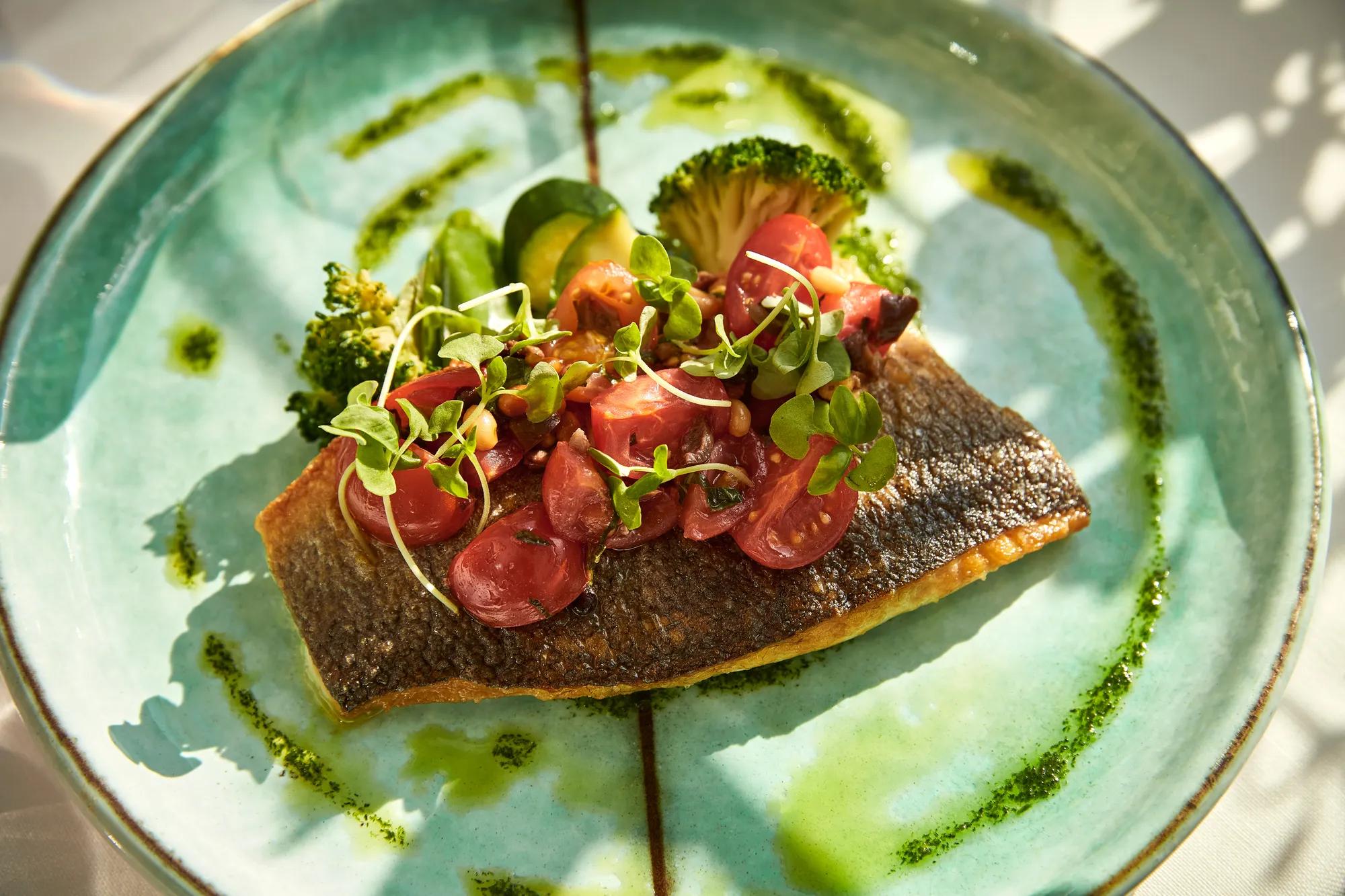Crispy-skinned salmon topped with cherry tomatoes and microgreens, served with broccoli and herb oil on a turquoise plate.