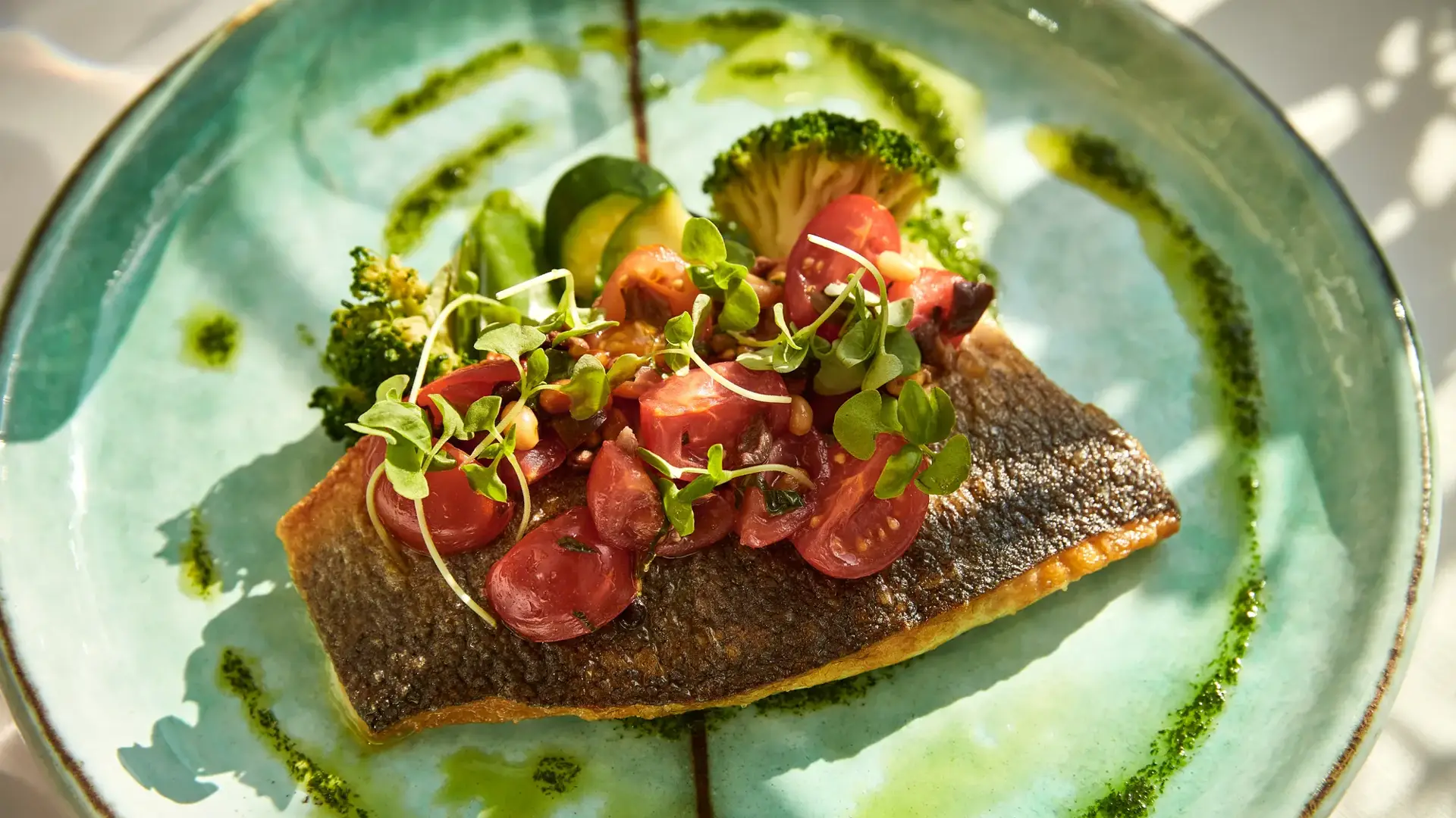 Crispy-skinned salmon topped with cherry tomatoes and microgreens, served with broccoli and herb oil on a turquoise plate.