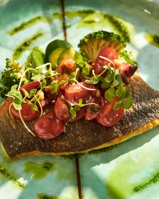 Crispy-skinned salmon topped with cherry tomatoes and microgreens, served with broccoli and herb oil on a turquoise plate.