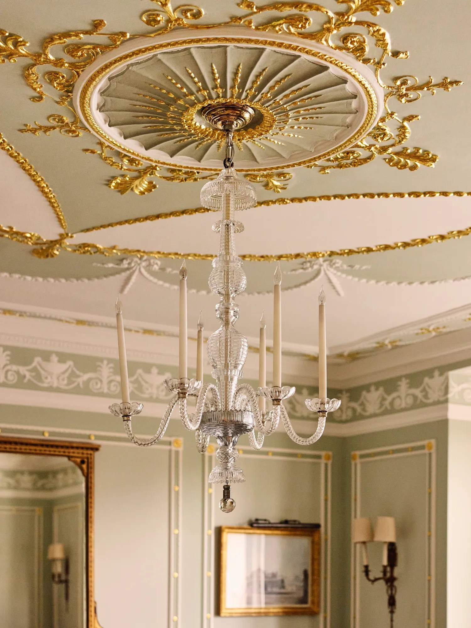 Glass chandelier with candle-style lights hanging from an ornate gold-detailed ceiling.