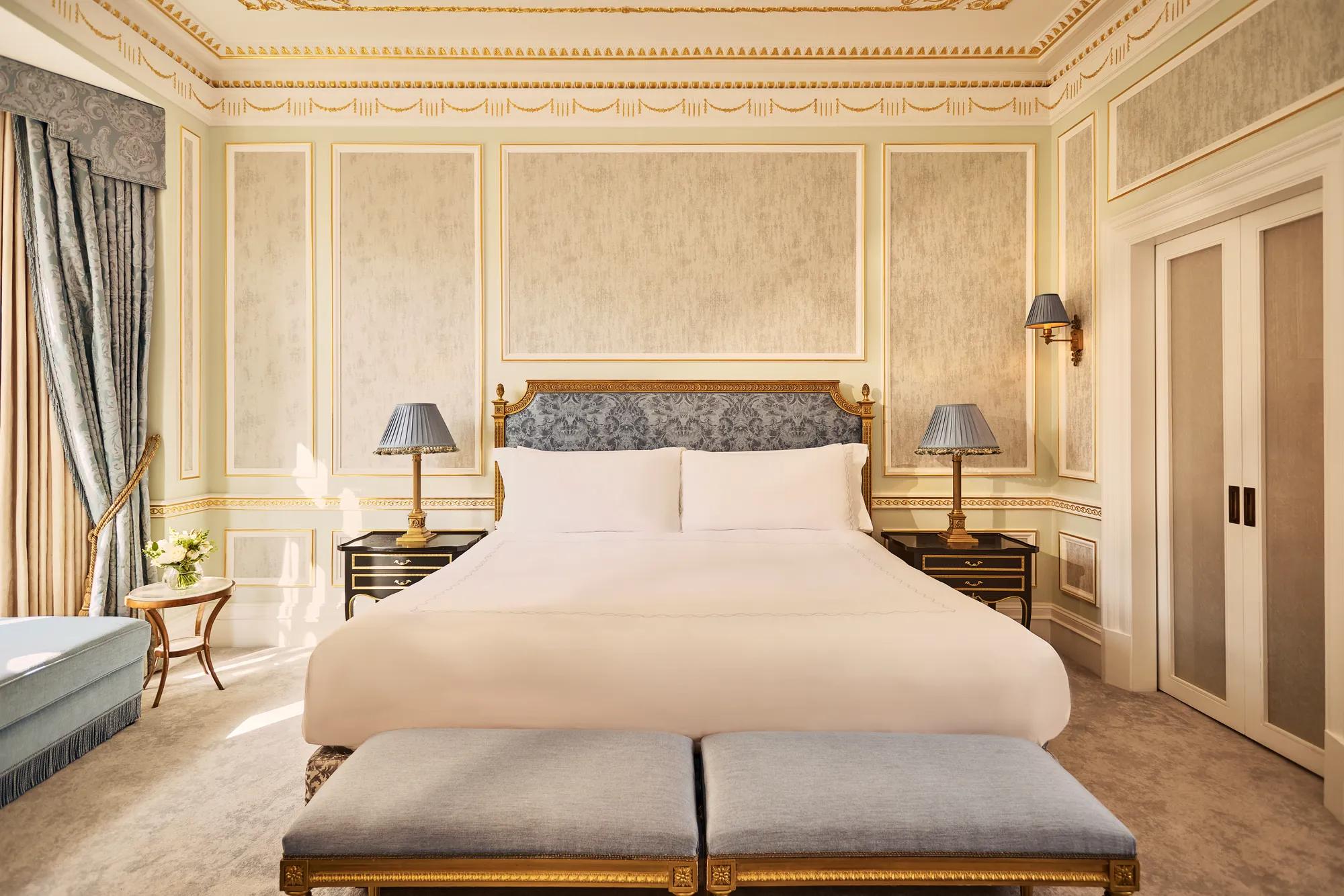 King-size bed with upholstered headboard in a classically styled bedroom featuring gold detailing, blue accents, and twin benches at the foot of the bed.