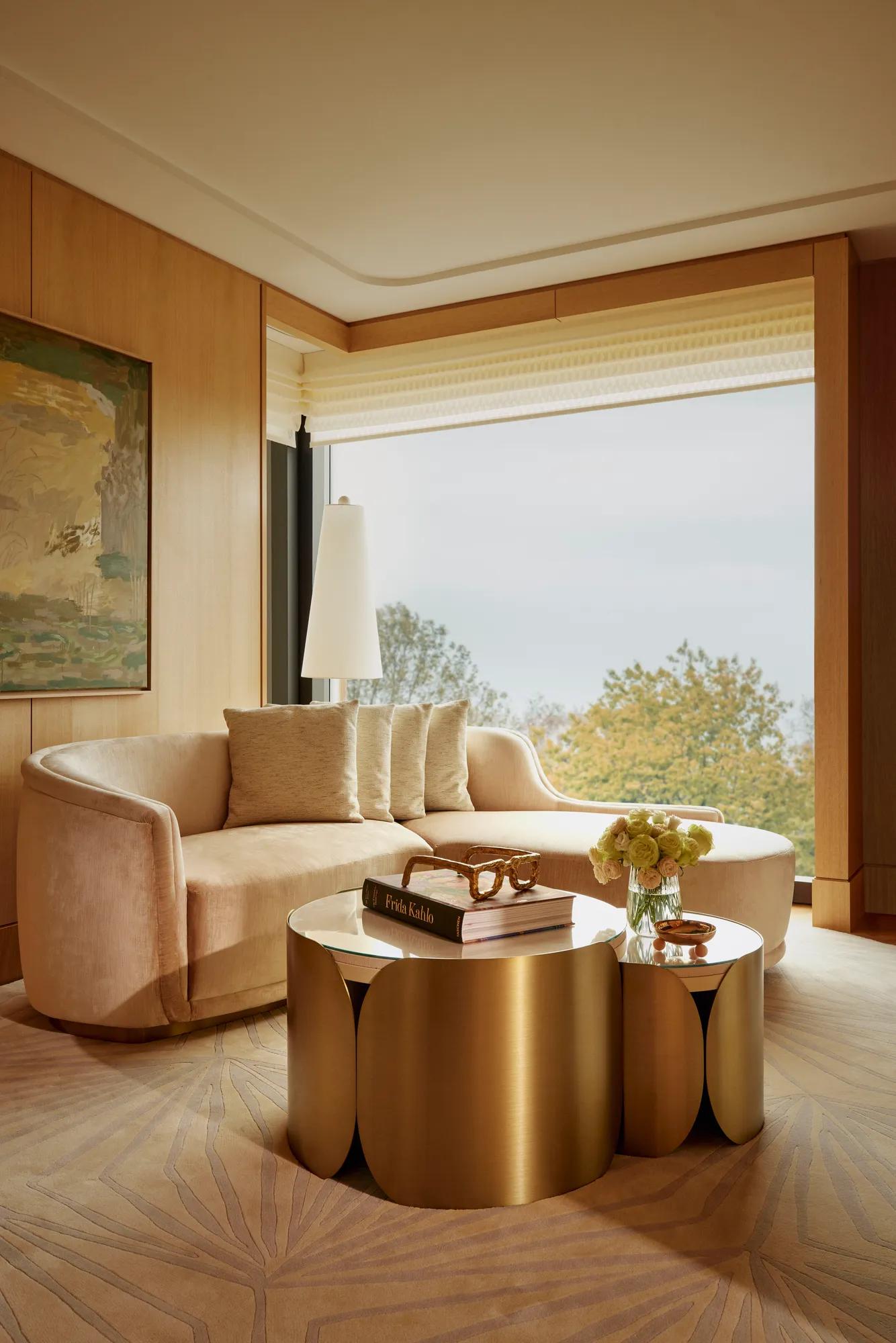 Curved cream sofa and gold coffee table beside full-height windows with leafy park views and modern artwork.