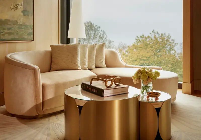 Curved cream sofa and gold coffee table beside full-height windows with leafy park views and modern artwork.