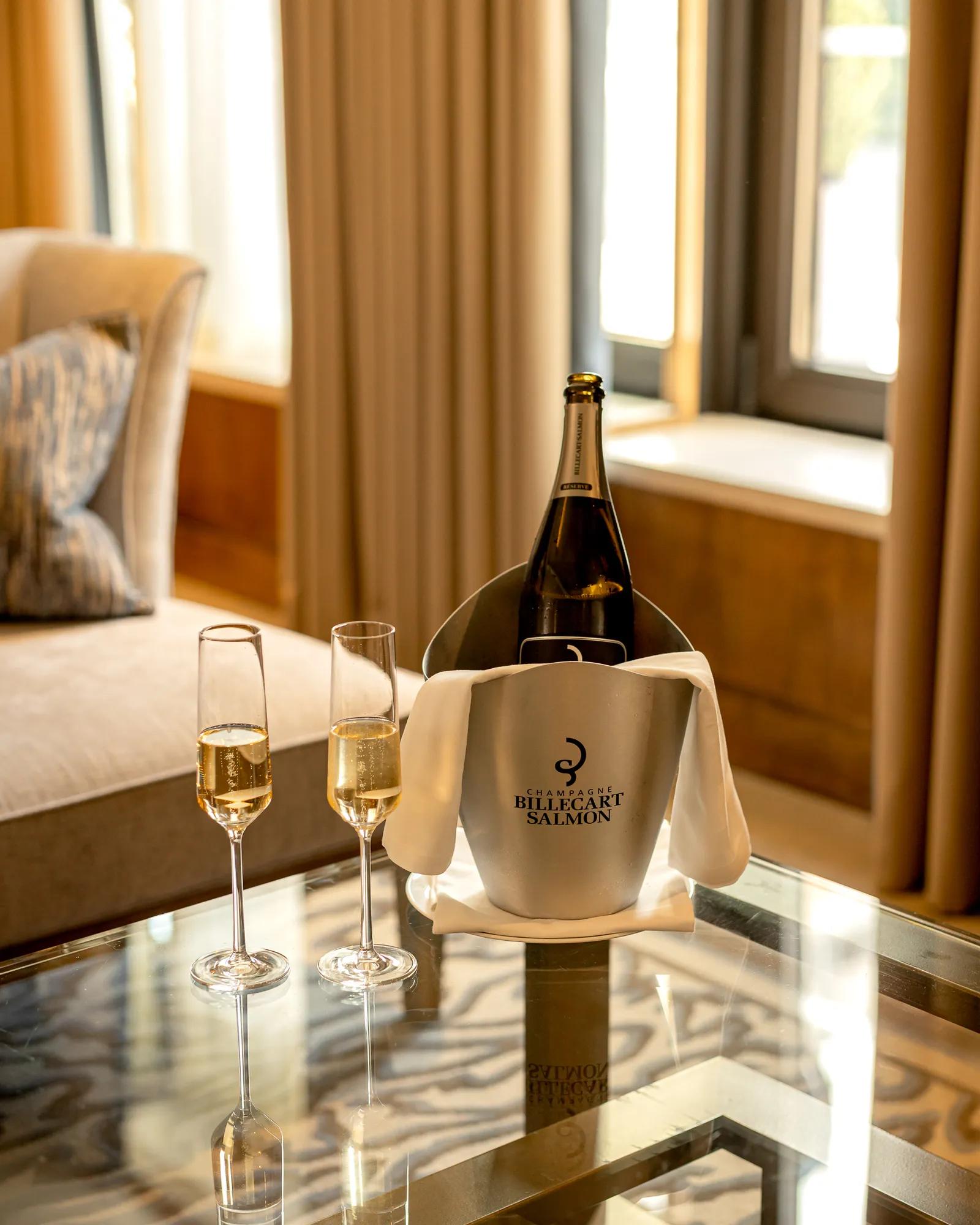 Champagne bottle in Billecart-Salmon ice bucket with two filled flutes on a glass table beside a sofa and window in a softly lit suite.
