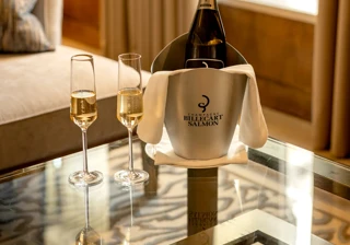 Champagne bottle in Billecart-Salmon ice bucket with two filled flutes on a glass table beside a sofa and window in a softly lit suite.