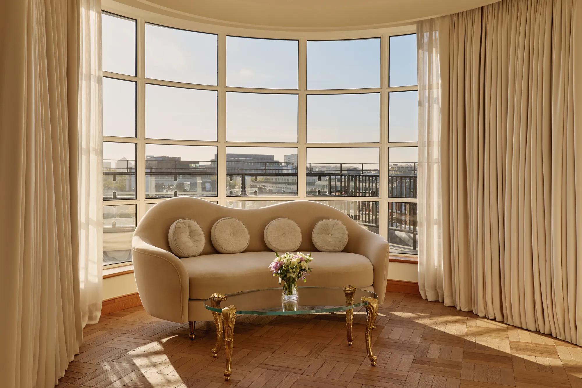 Curved sitting area with a cream sofa and circular cushions beside a glass coffee table with gold legs, framed by floor-to-ceiling windows offering panoramic city views.