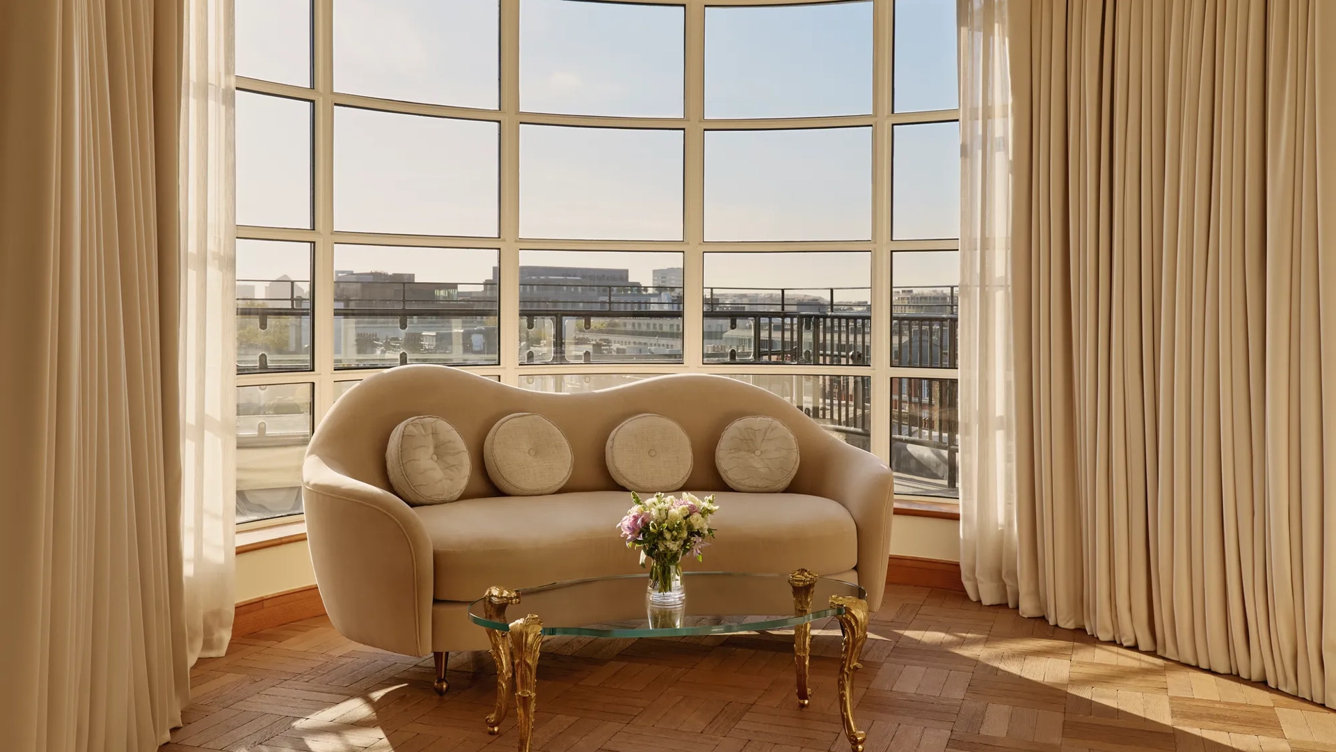 Curved sitting area with a cream sofa and circular cushions beside a glass coffee table with gold legs, framed by floor-to-ceiling windows offering panoramic city views.