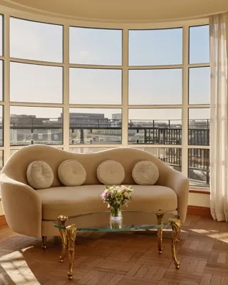 Curved sitting area with a cream sofa and circular cushions beside a glass coffee table with gold legs, framed by floor-to-ceiling windows offering panoramic city views.
