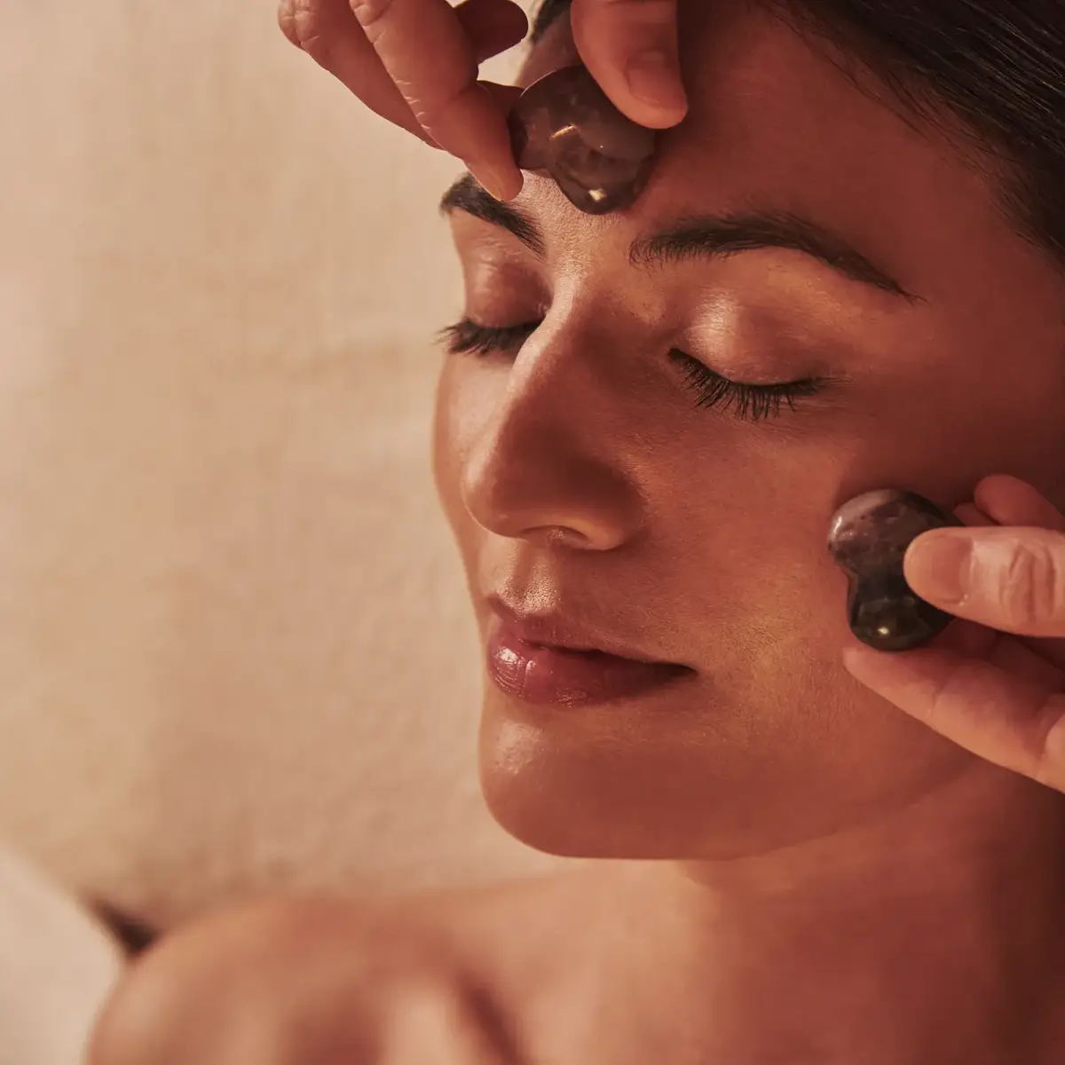 Person receiving a facial massage with smooth stones applied to the forehead and cheek while lying relaxed.