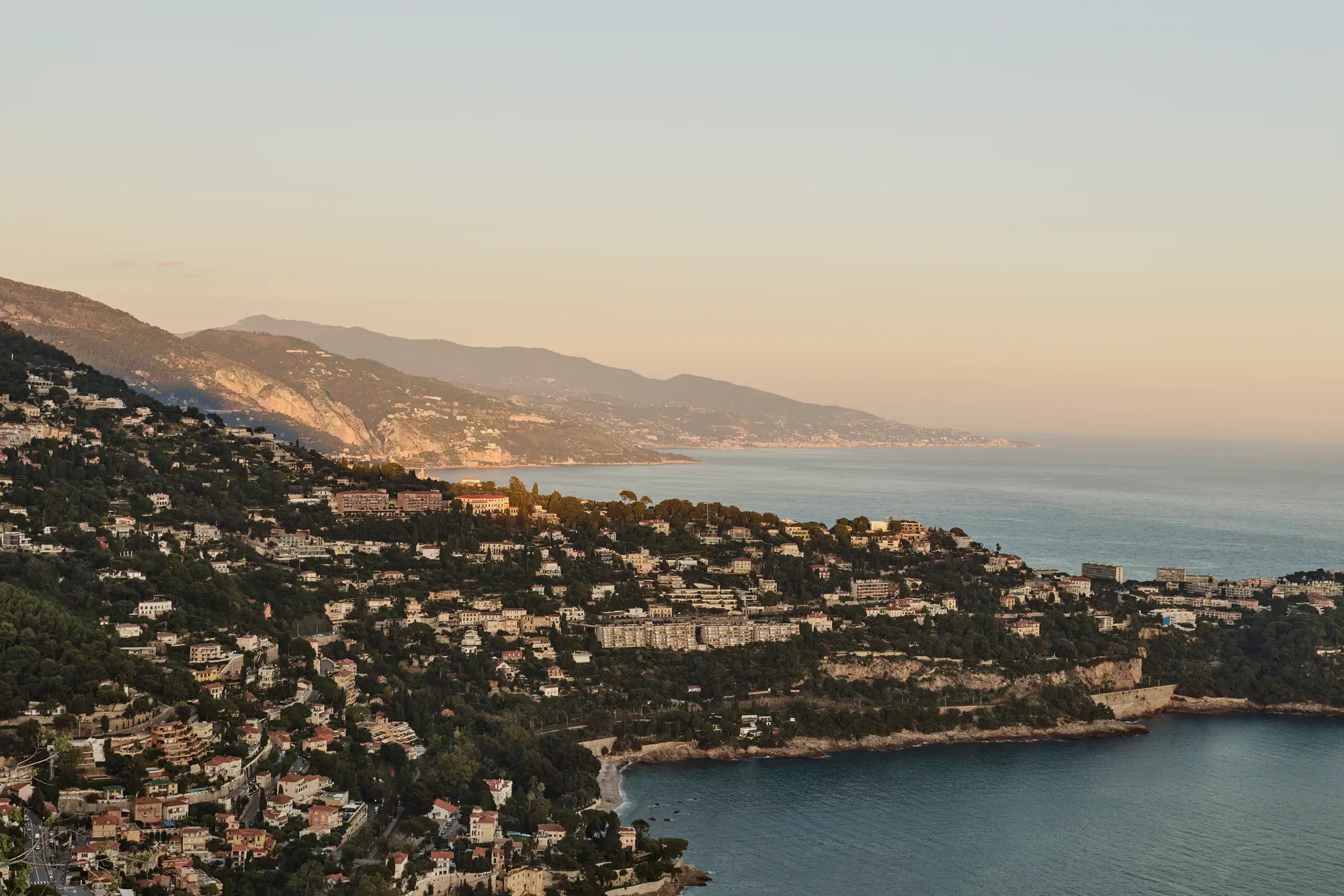 Golden-hour view of coastal hillside town with villas, cliffs and calm sea stretching along the Riviera coastline.