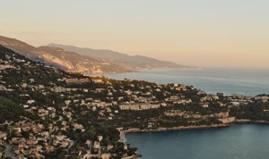 Golden-hour view of coastal hillside town with villas, cliffs and calm sea stretching along the Riviera coastline.