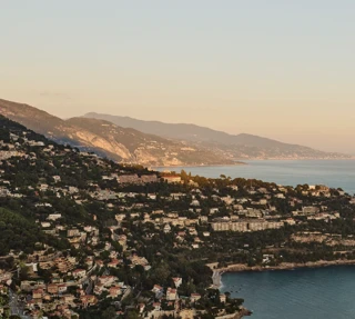Golden-hour view of coastal hillside town with villas, cliffs and calm sea stretching along the Riviera coastline.