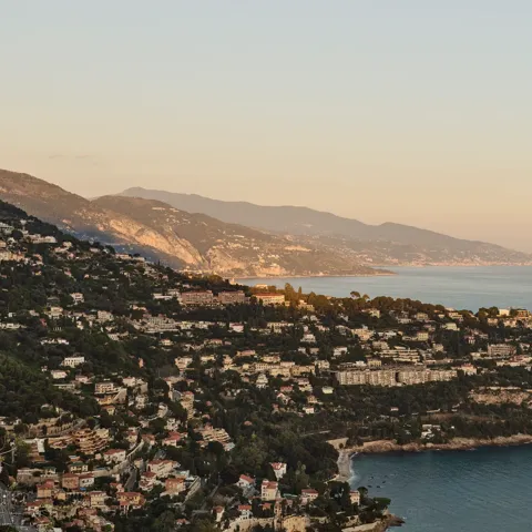 Golden-hour view of coastal hillside town with villas, cliffs and calm sea stretching along the Riviera coastline.