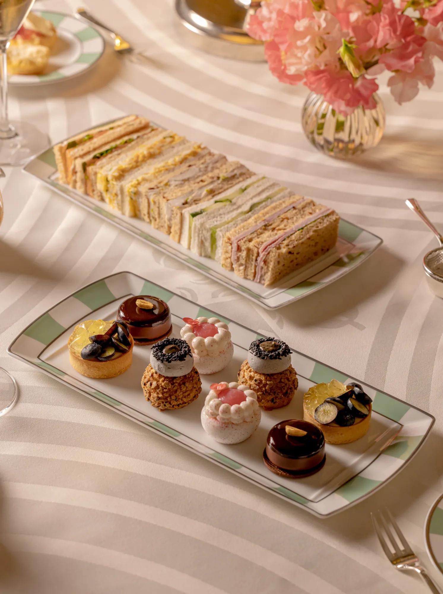 Tiered afternoon tea selection on a striped tablecloth, featuring neatly stacked finger sandwiches and assorted miniature pastries beside a vase of pink flowers.