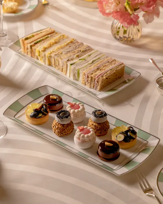 Tiered afternoon tea selection on a striped tablecloth, featuring neatly stacked finger sandwiches and assorted miniature pastries beside a vase of pink flowers.