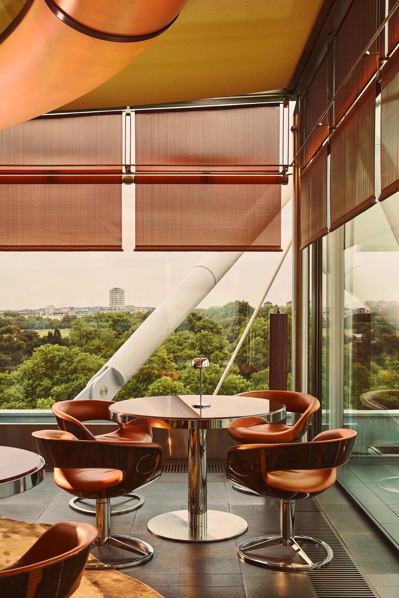 Floor-to-ceiling windows frame a leafy park view beside a terrace table with copper-toned chairs and contemporary metal detailing.