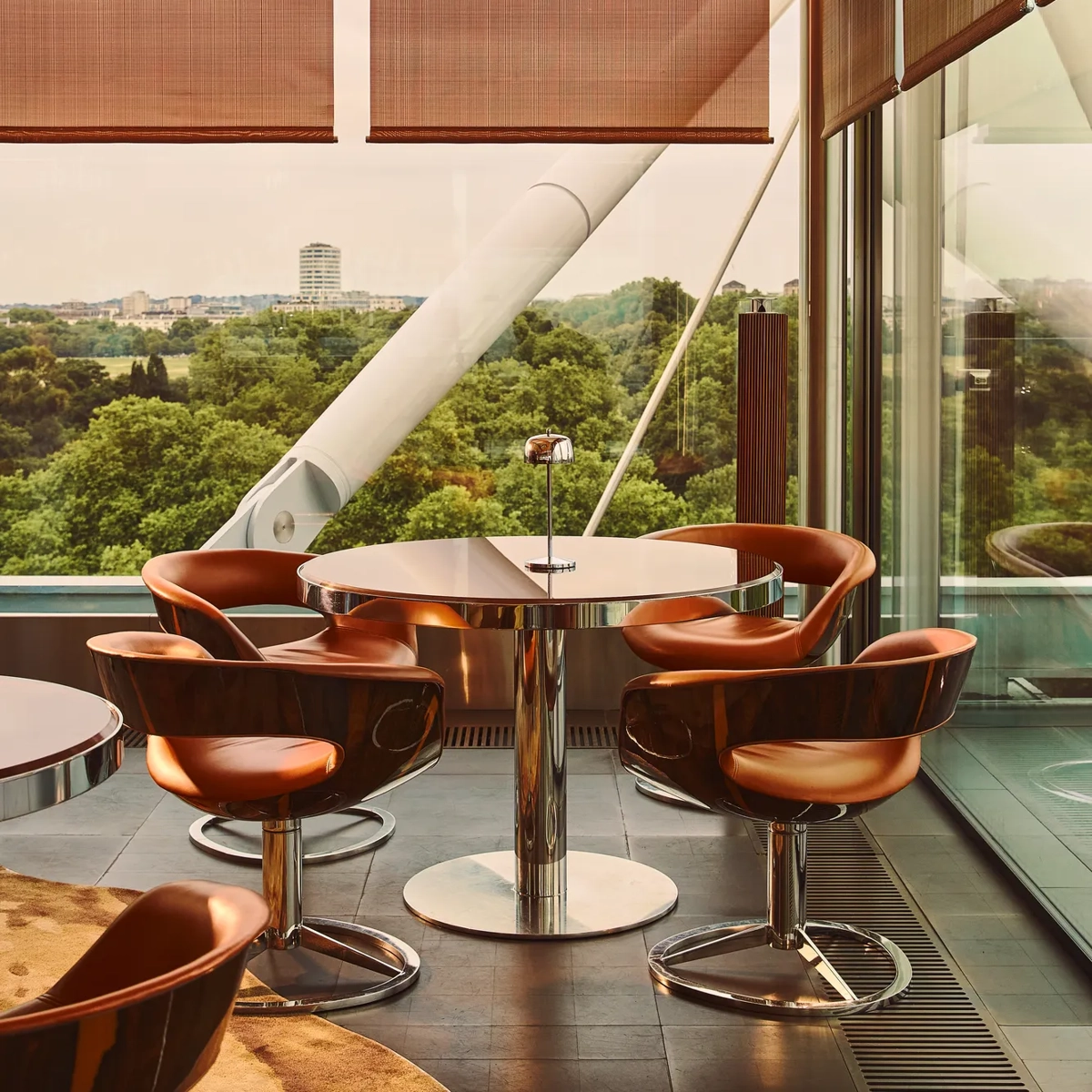 Floor-to-ceiling windows frame a leafy park view beside a terrace table with copper-toned chairs and contemporary metal detailing.