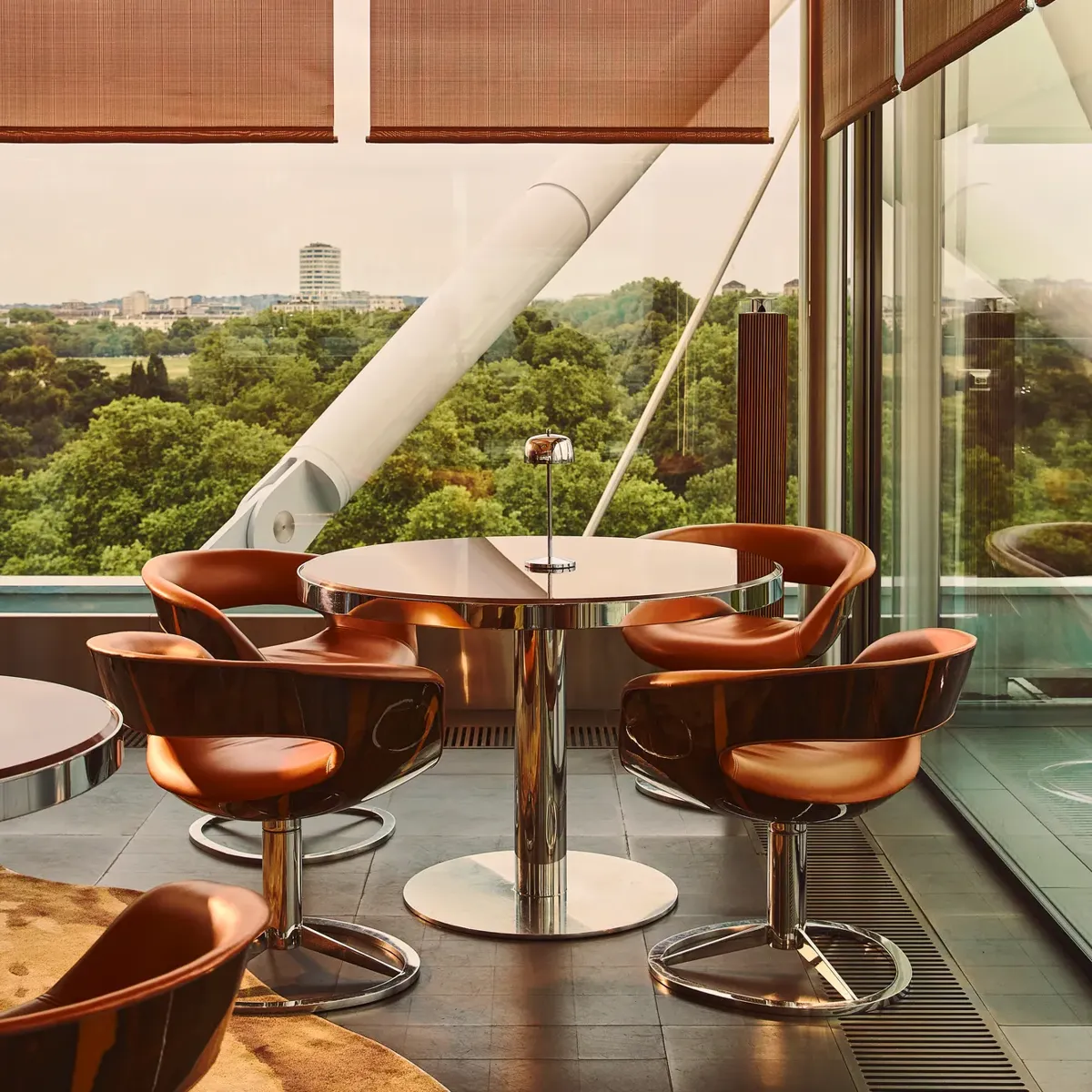 Floor-to-ceiling windows frame a leafy park view beside a terrace table with copper-toned chairs and contemporary metal detailing.