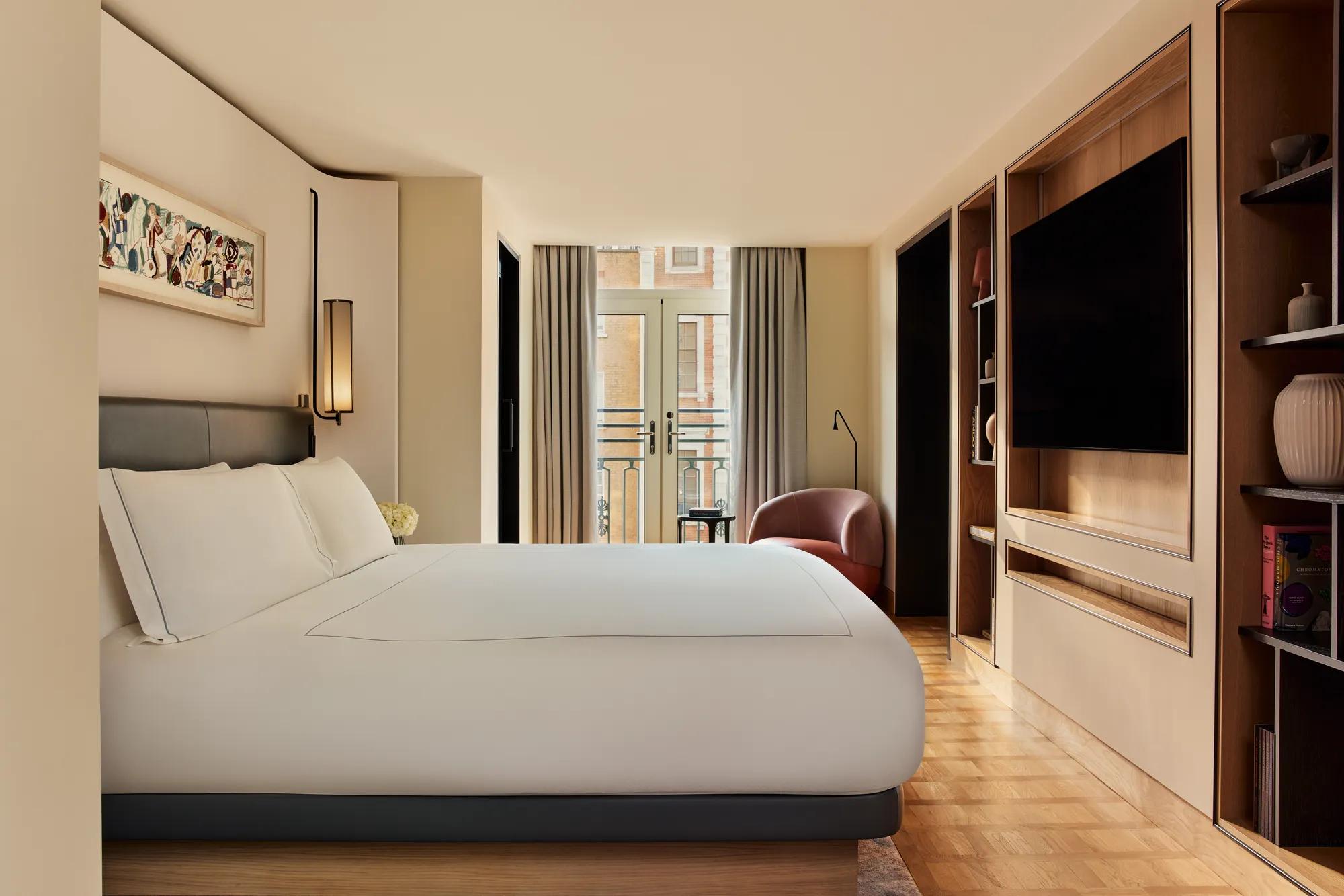 Spacious bedroom in the Knightsbridge Suite with a large bed in white linens, built-in shelving with a flat-screen TV, and French doors opening onto a balcony with city views.