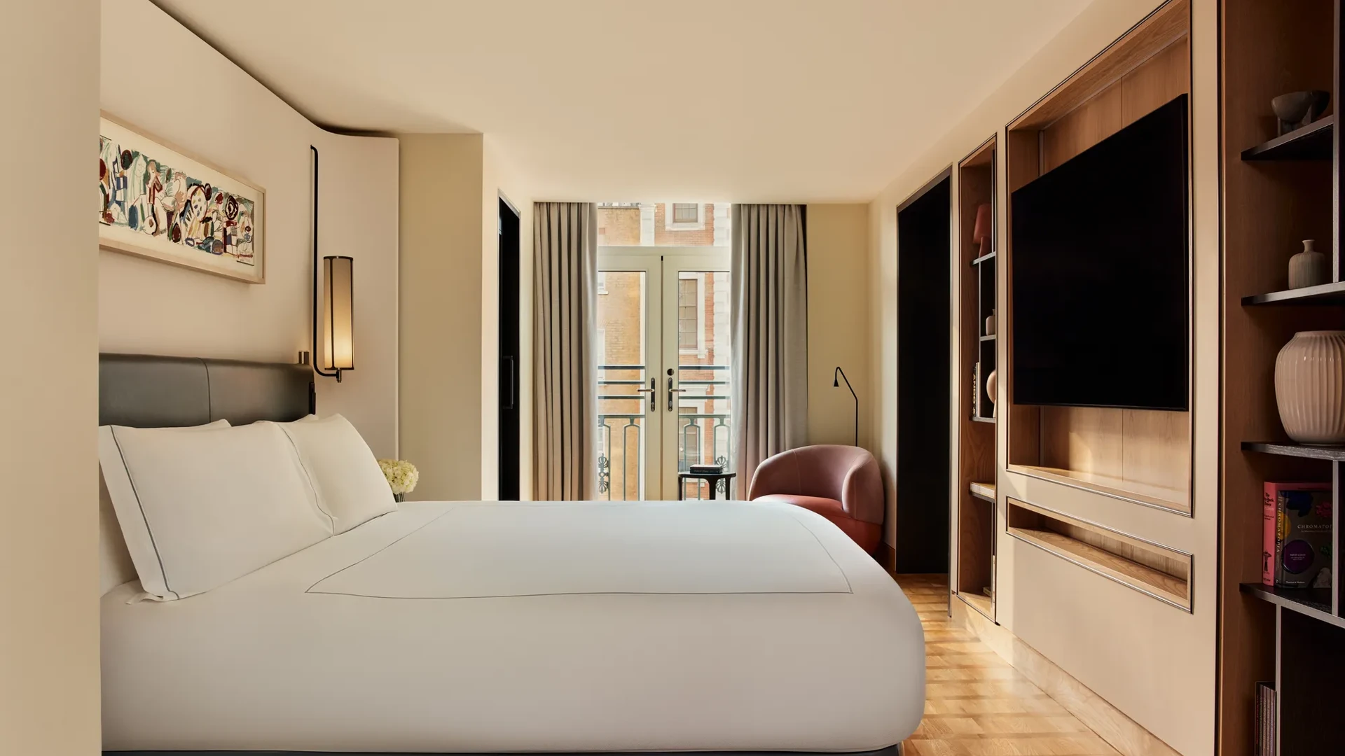 Spacious bedroom in the Knightsbridge Suite with a large bed in white linens, built-in shelving with a flat-screen TV, and French doors opening onto a balcony with city views.