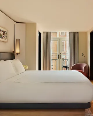 Spacious bedroom in the Knightsbridge Suite with a large bed in white linens, built-in shelving with a flat-screen TV, and French doors opening onto a balcony with city views.