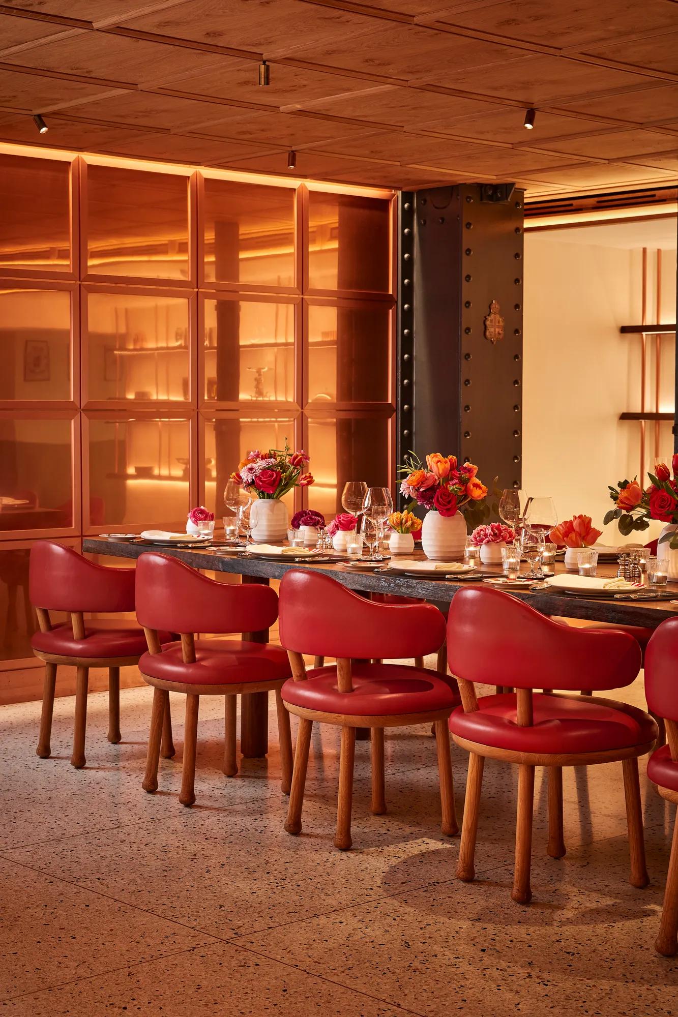Contemporary dining area featuring a long wooden table surrounded by red leather chairs. The table is elegantly set with glassware, candles, and vibrant floral arrangements, framed by warm amber lighting and modern architectural details.
