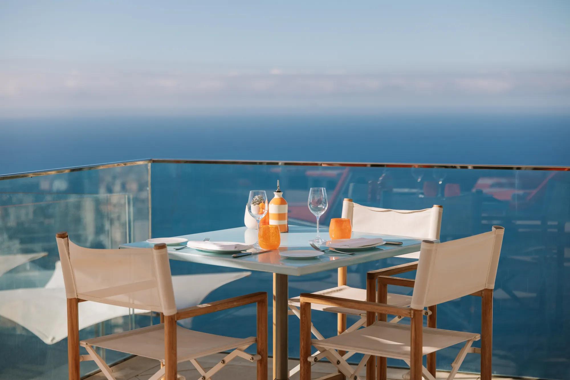 Table de terrasse en bord de mer dressée pour deux avec chaises blanches et vue sur l’océan.