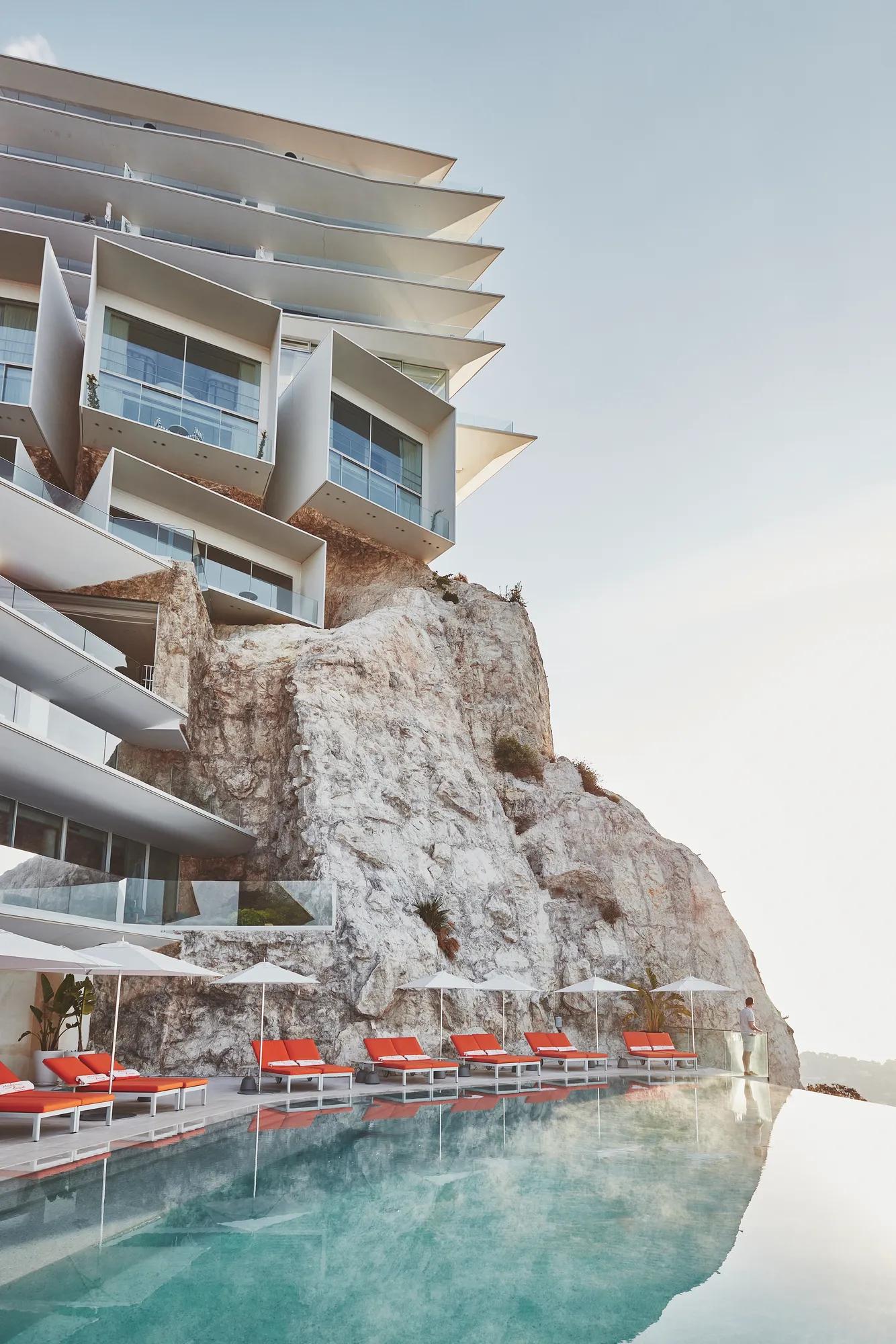 Modern cliffside resort with an infinity pool, red loungers, and dramatic rock formations.