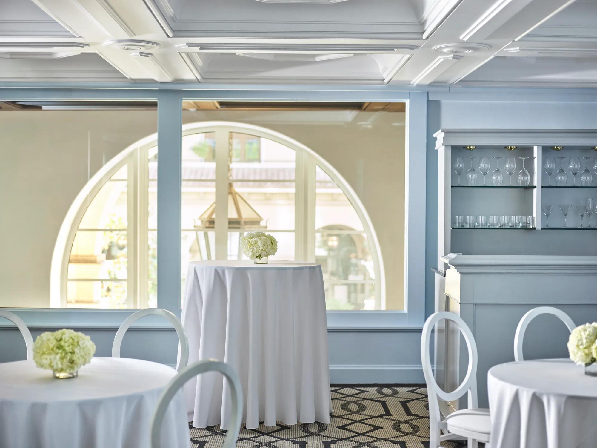 Wilshire Room reception setup with white highboy tables, round-back chairs, pale blue paneling, and an arched window beside a built-in glassware bar.