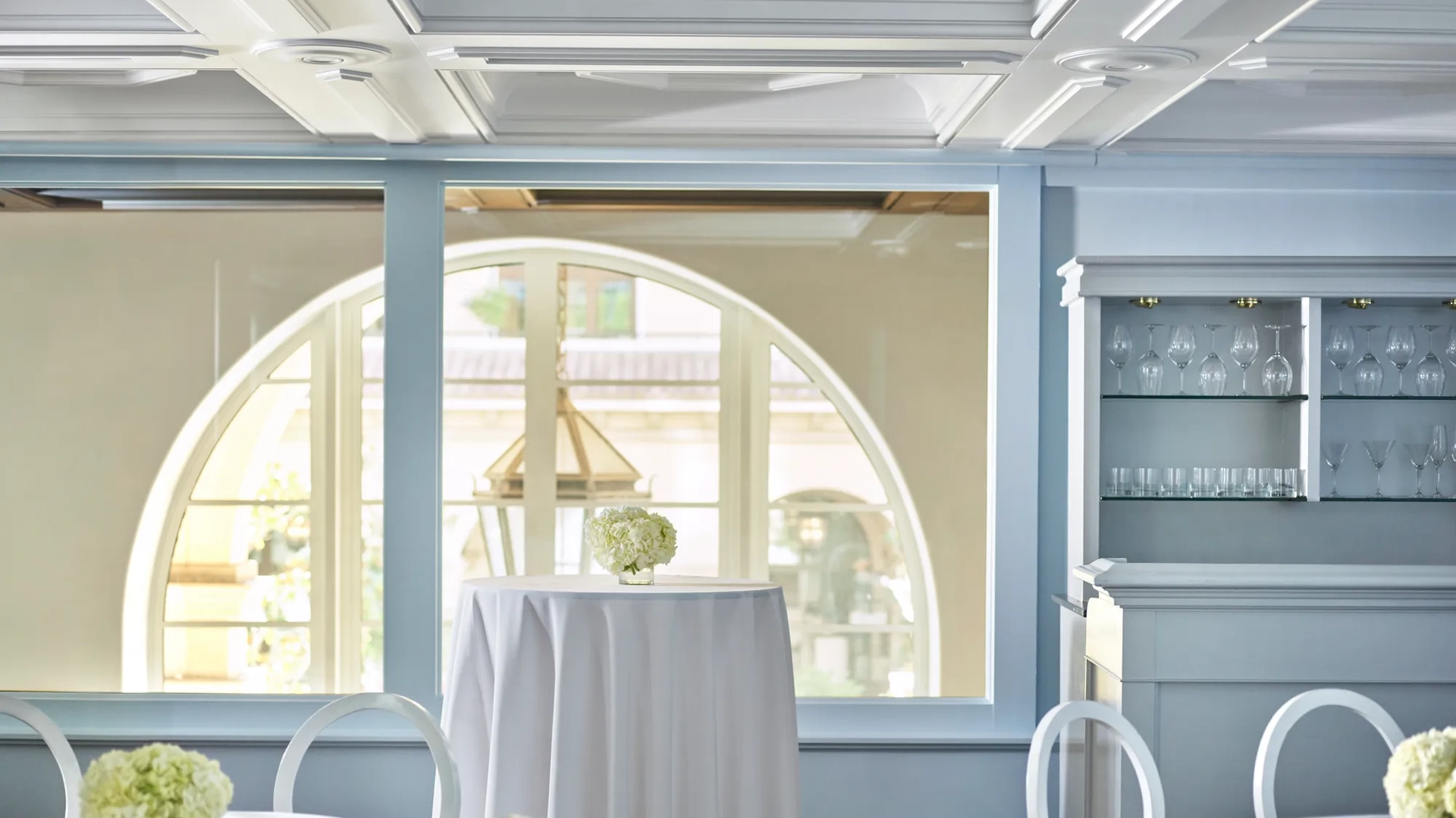 Wilshire Room reception setup with white highboy tables, round-back chairs, pale blue paneling, and an arched window beside a built-in glassware bar.