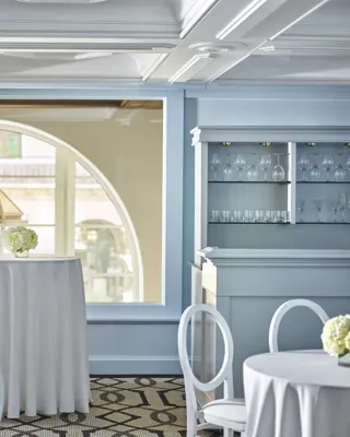 Wilshire Room reception setup with white highboy tables, round-back chairs, pale blue paneling, and an arched window beside a built-in glassware bar.