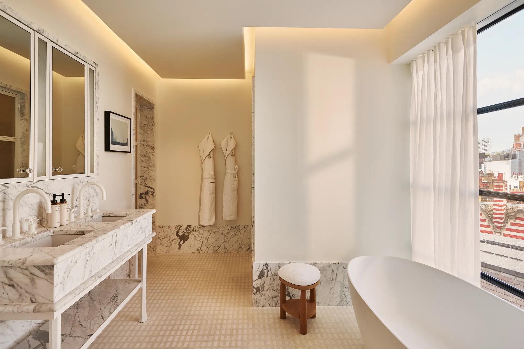 Light-filled bathroom with marble vanity and mirrors, freestanding bathtub by window, robes on wall, and city view.