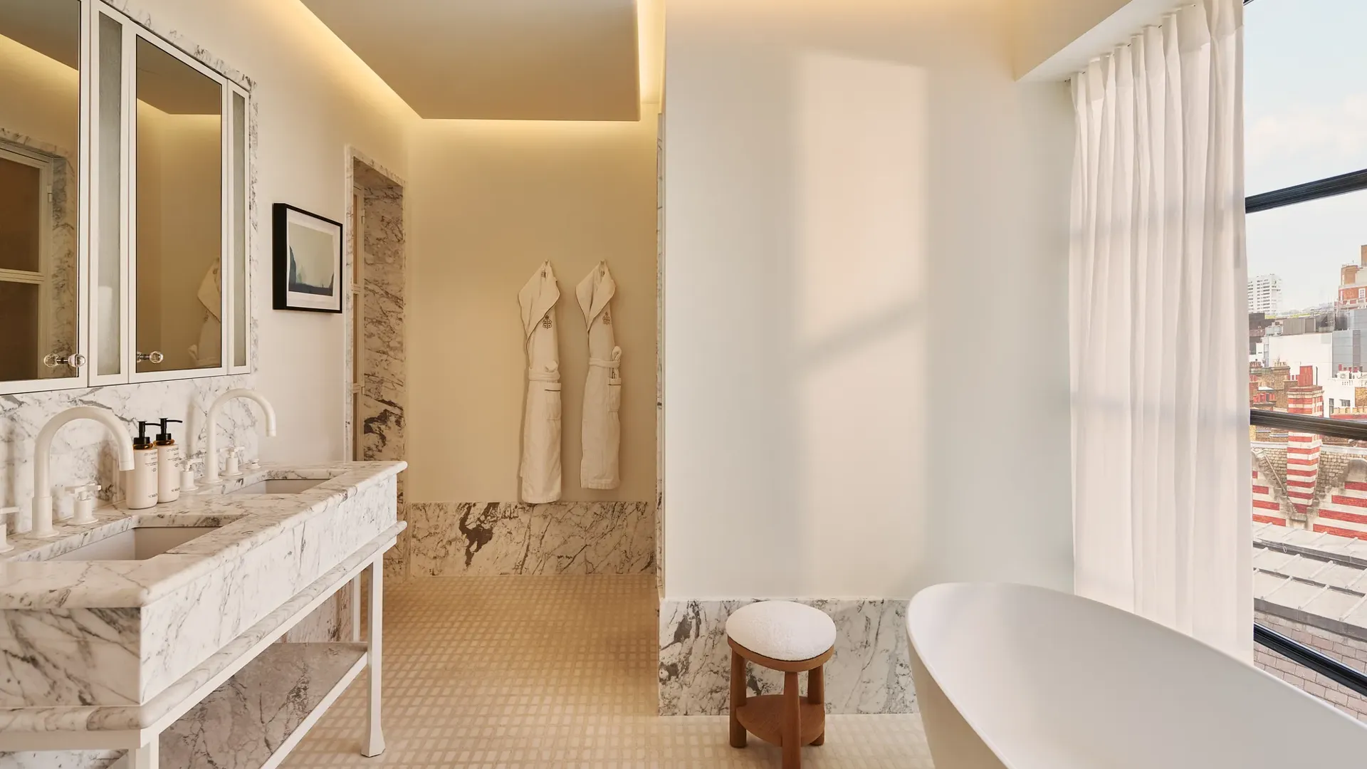 Light-filled bathroom with marble vanity and mirrors, freestanding bathtub by window, robes on wall, and city view.