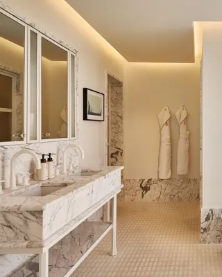 Light-filled bathroom with marble vanity and mirrors, freestanding bathtub by window, robes on wall, and city view.