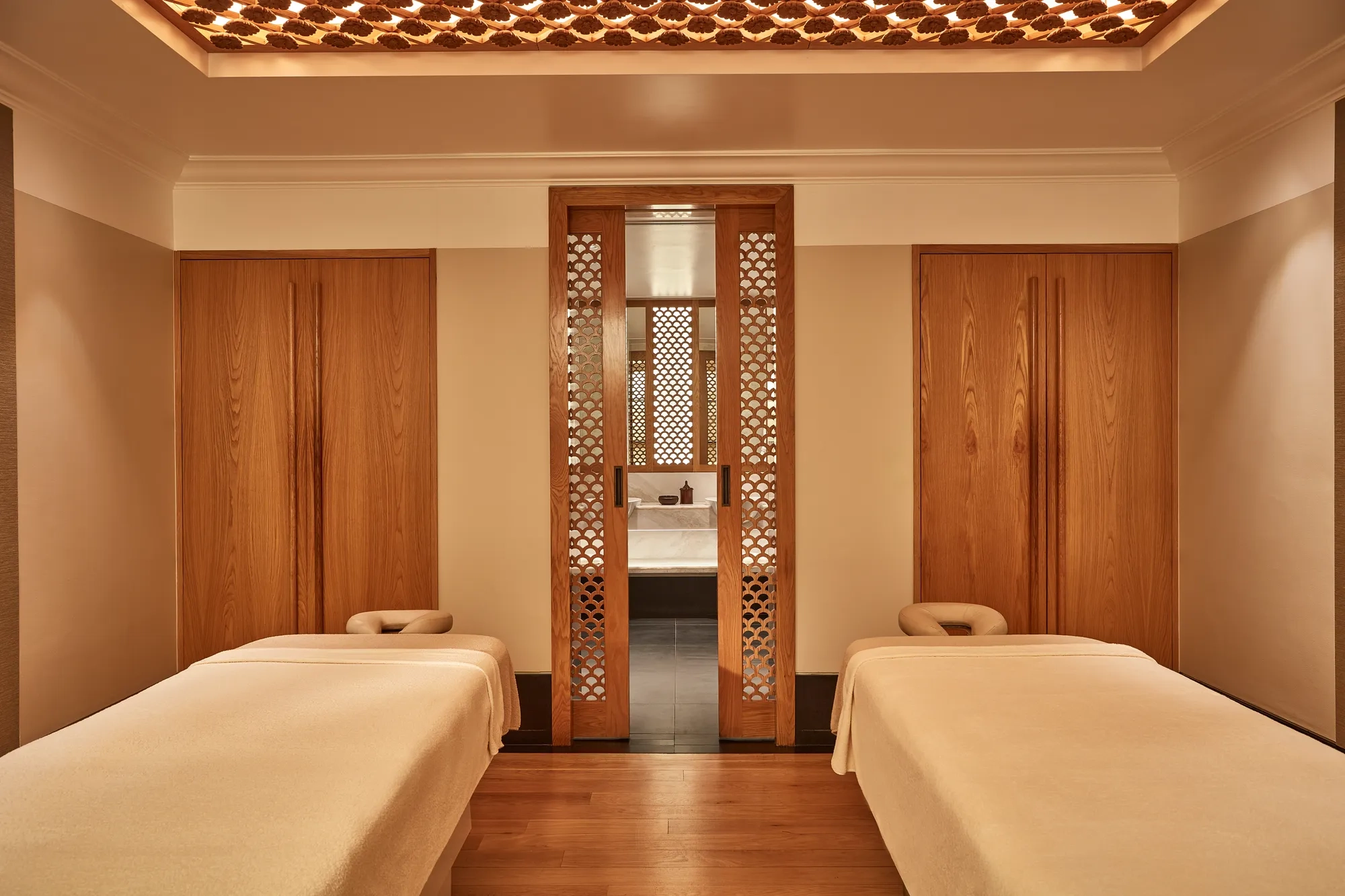 Calm spa treatment room with two massage tables, wooden lattice doors, and warm soft lighting.